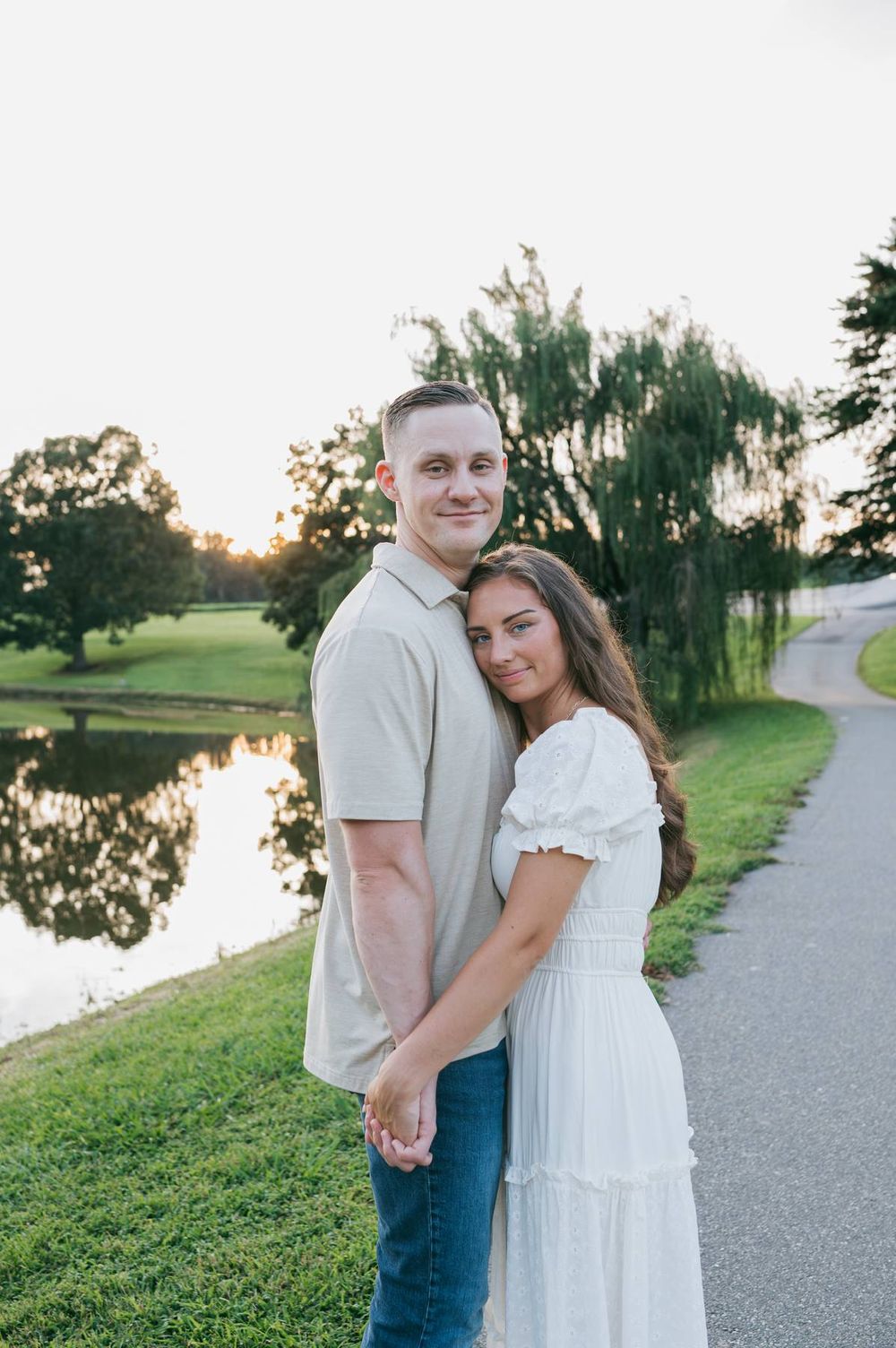 Smiling couple holding hands and posing together beside a tranquil pond at golden hour, engagement photographer Mooresville NC
