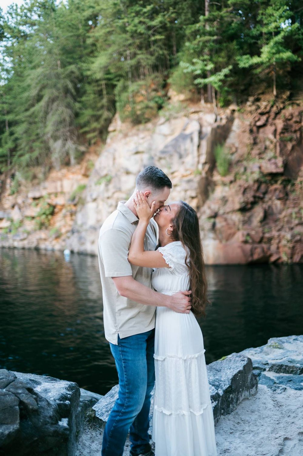 Couple sharing a romantic kiss on rocky shoreline with dramatic cliffs and dark water behind, engagement photographer Mooresville NC