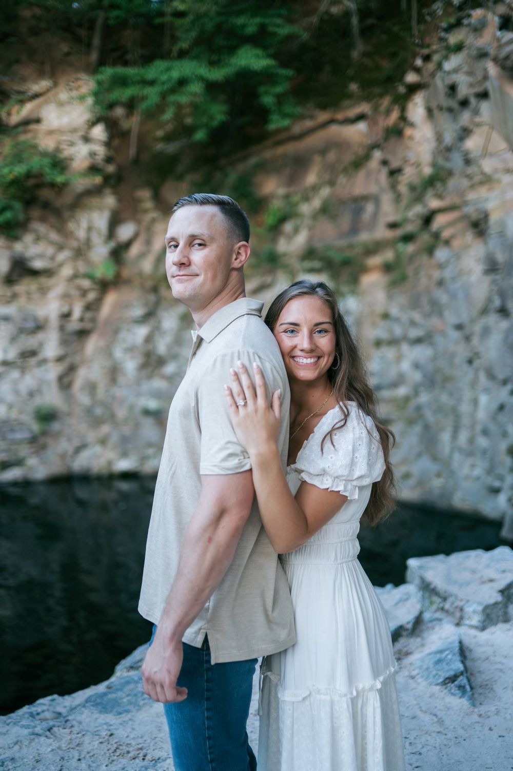 Couple posing back-to-back at rocky waterfall, woman in white dress hugging man in beige shirt, smiling outdoors, engagement photographer NC