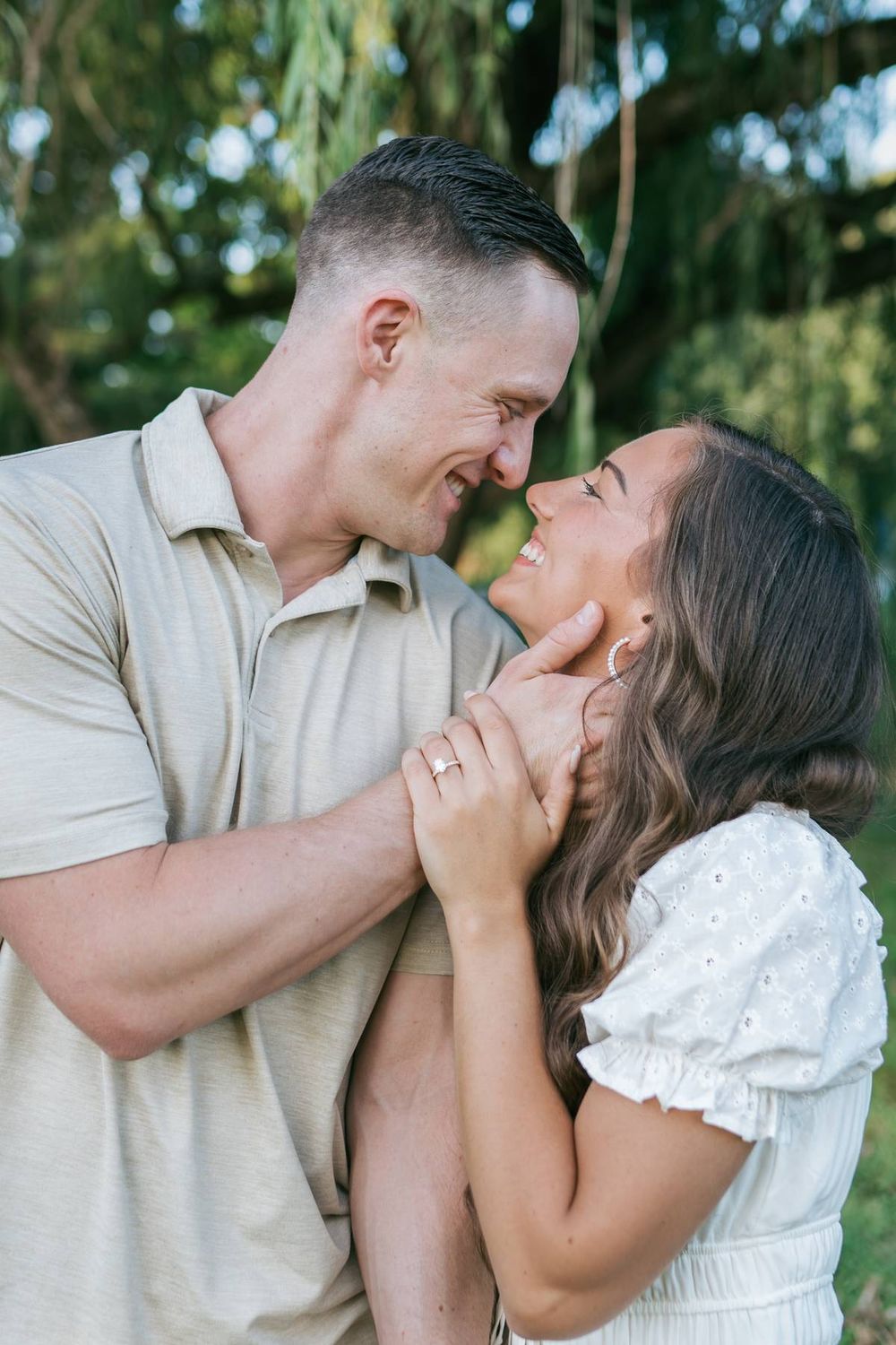 Happy engaged couple nearly kissing under willow tree, woman showing engagement ring while man holds her face tenderly, photographer NC