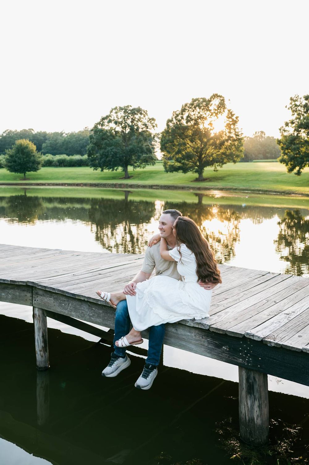 Couple sitting on wooden dock at sunset, woman in white dress embracing man by serene reflective lake, engagement photographer Greensboro NC