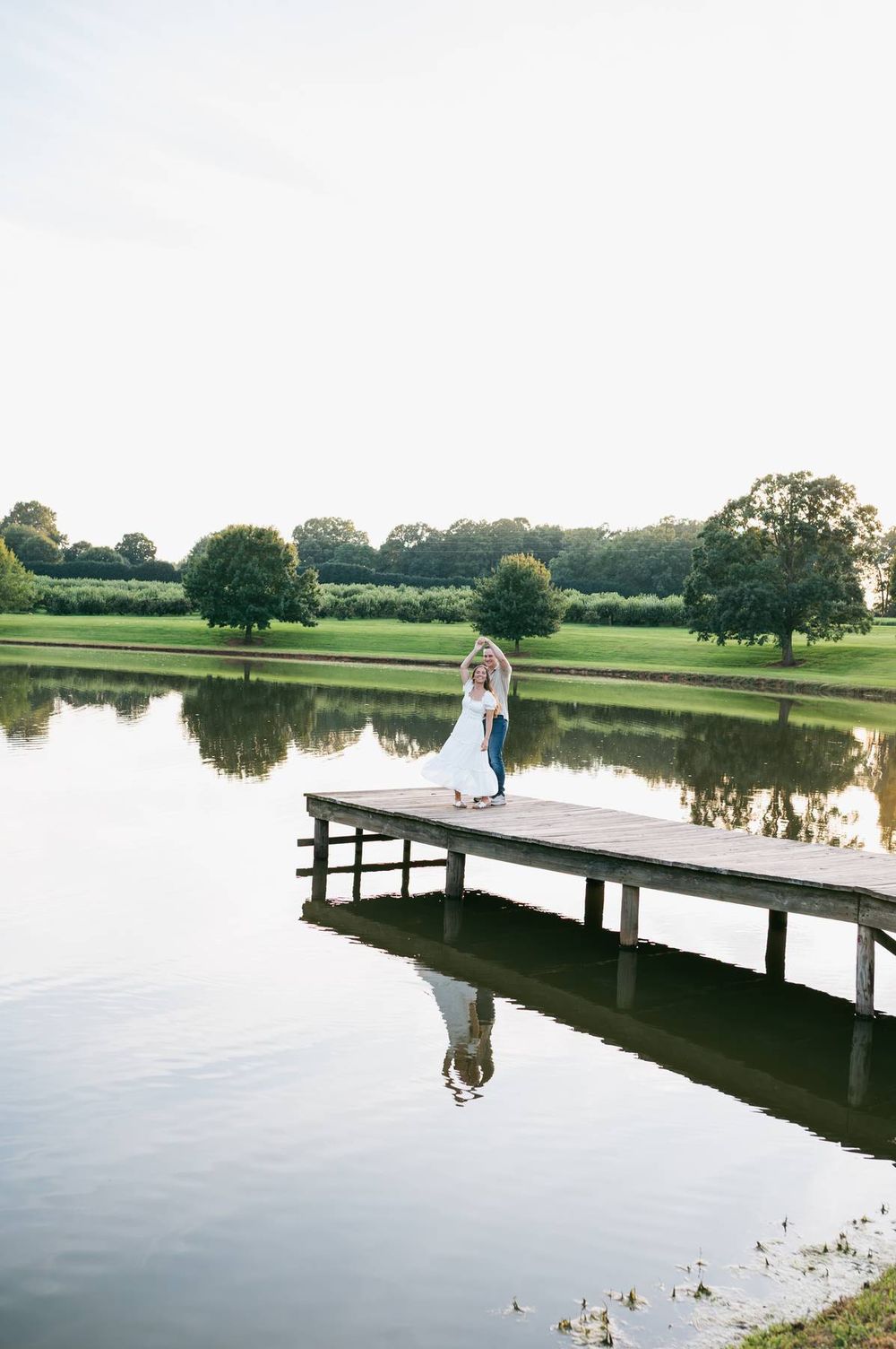 Engaged couple dancing on wooden dock over calm lake at golden hour, surrounded by lush green landscape, engagement photographer Asheboro NC