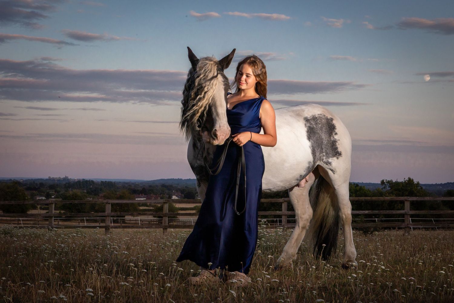 Woman in blue gown poses with gray horse at sunset in a field.