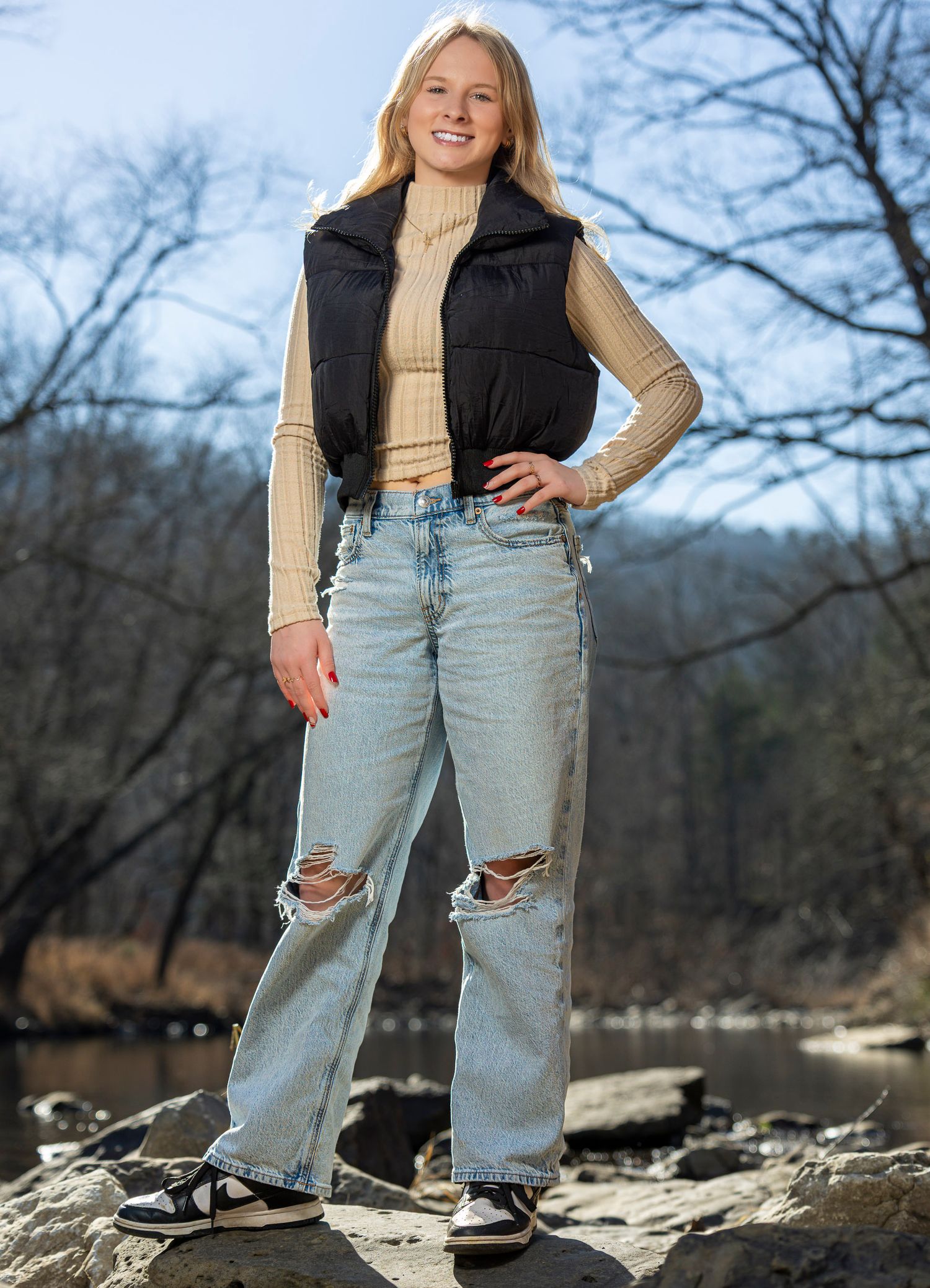 Smiling young blonde woman in black puffer vest and cream sweater sitting outdoors with arms crossed.
