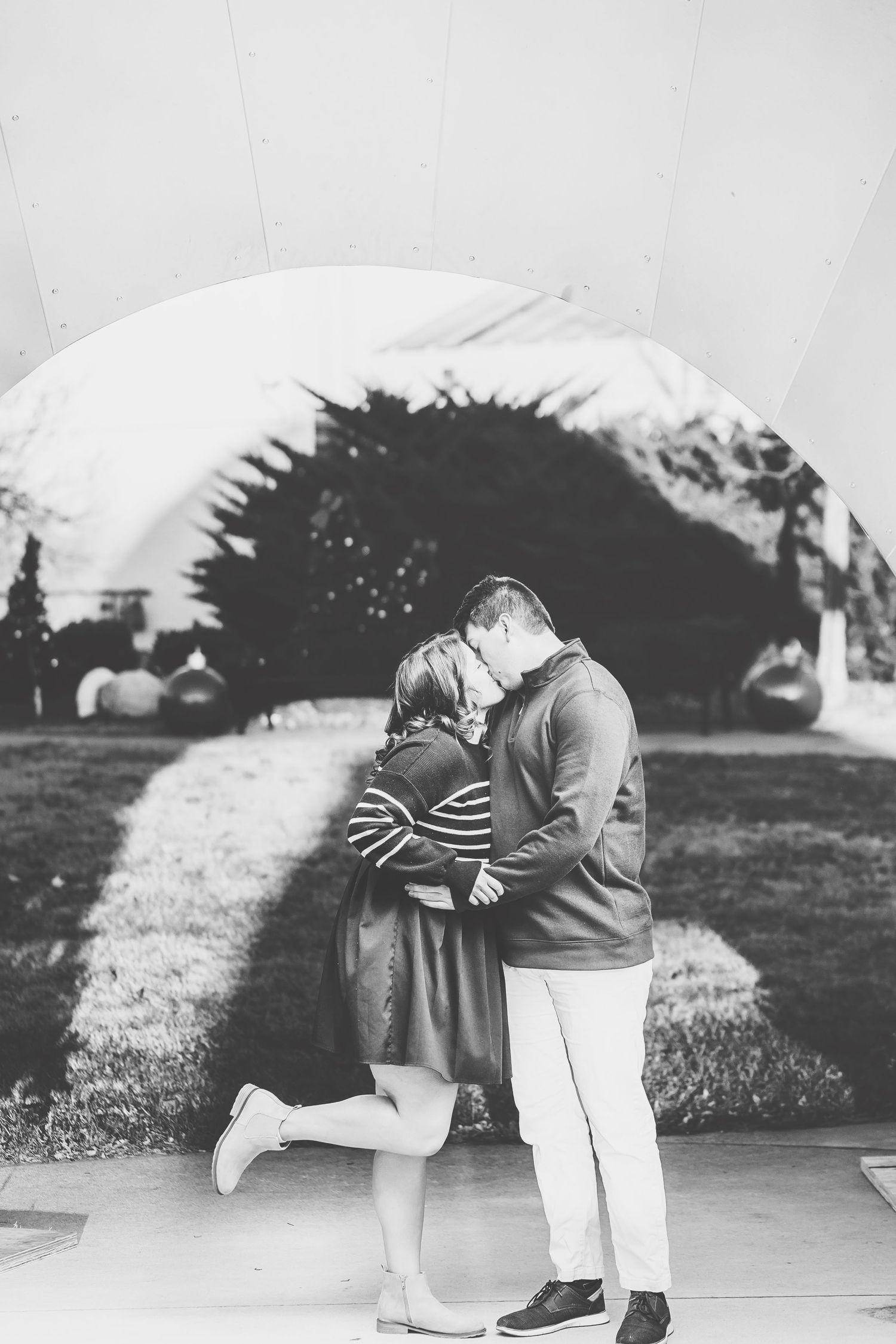 Black and white photo of a couple sharing a romantic kiss outdoors under an arch, woman lifting one leg.