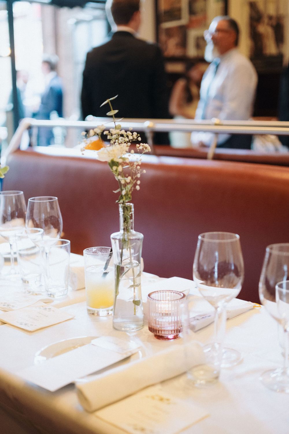 Elegant restaurant table setting with wine glasses, floral centerpiece, and candles, with guests mingling in background.