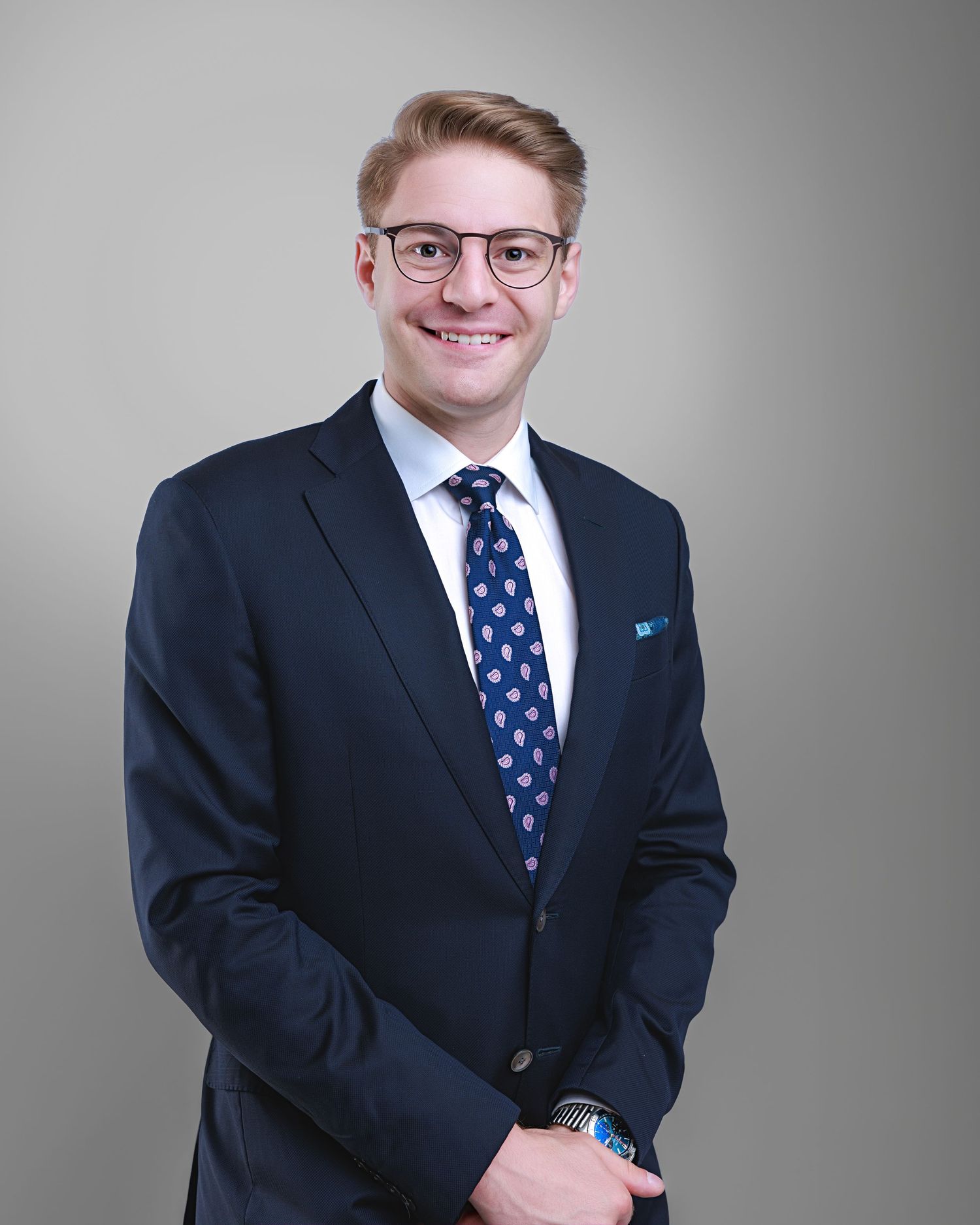 Young professional man in navy suit and blue tie smiling confidently for a corporate headshot photo.