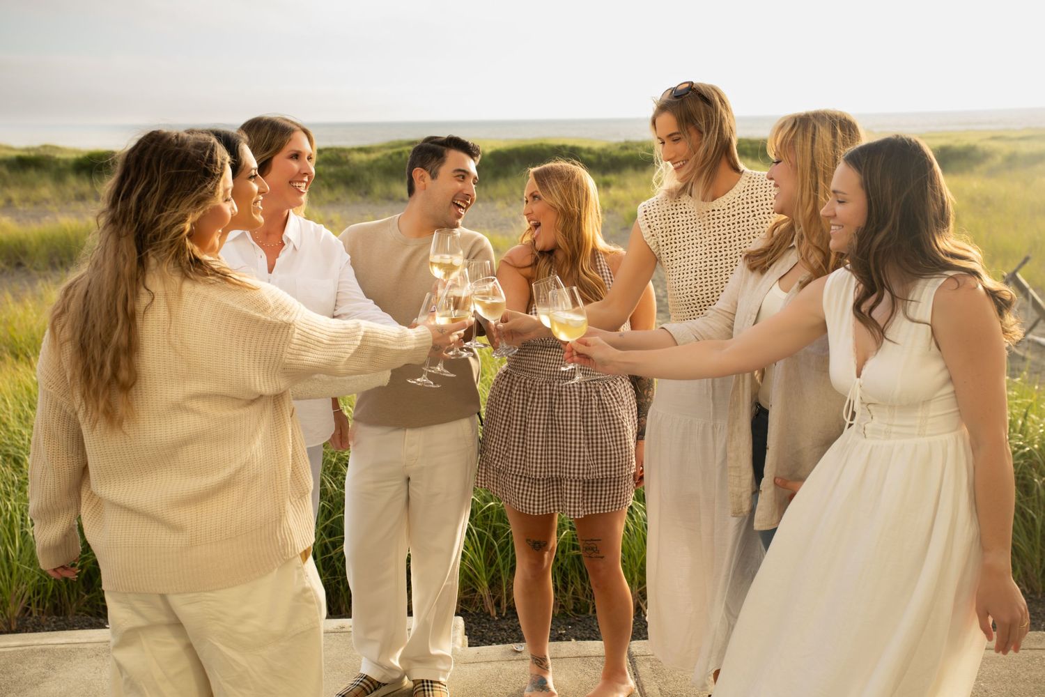 Group of friends in white outfits toasting with champagne glasses outdoors at a summer celebration.