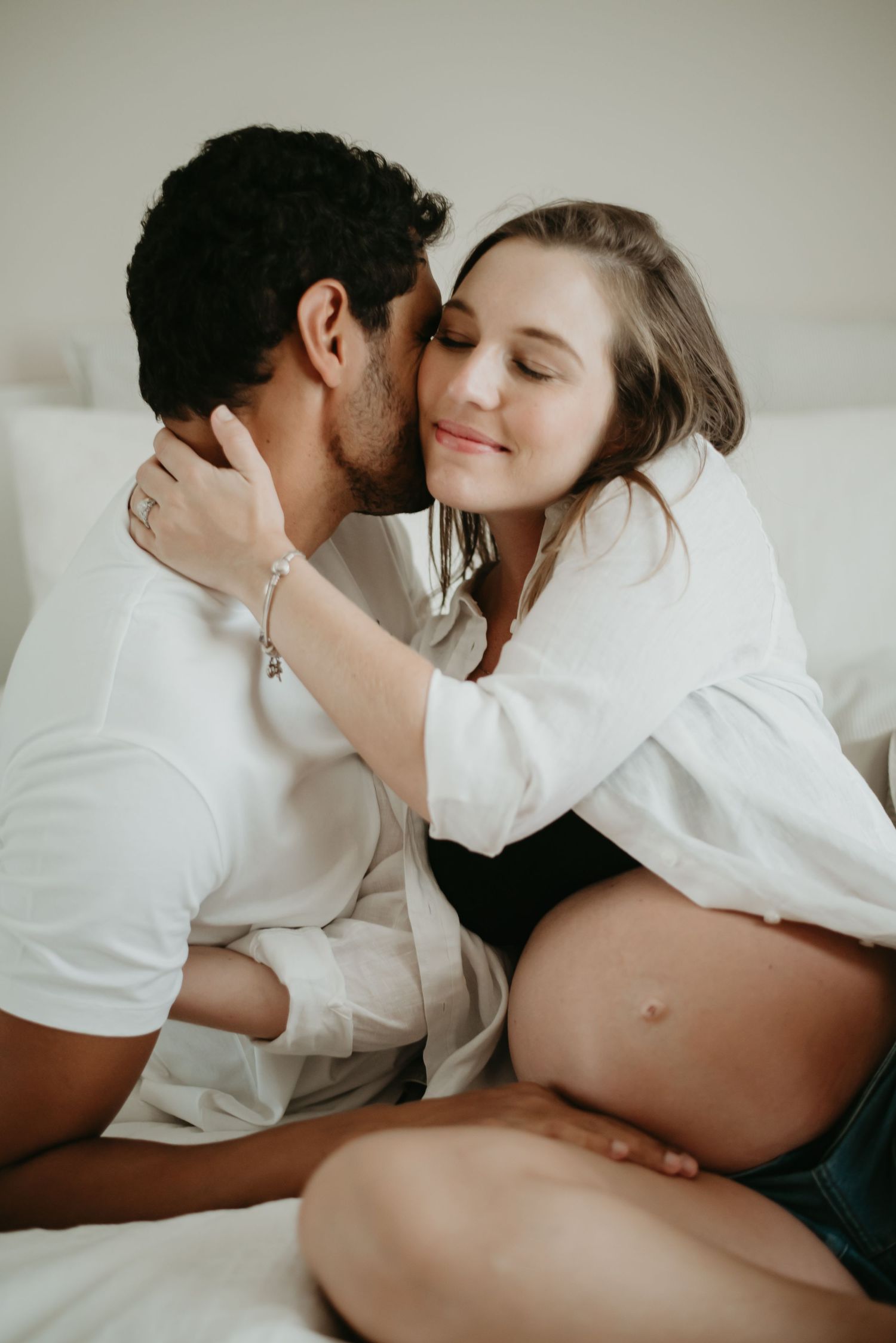 A pregnant woman smiles as her partner kisses her cheek while sitting together on a white bed.