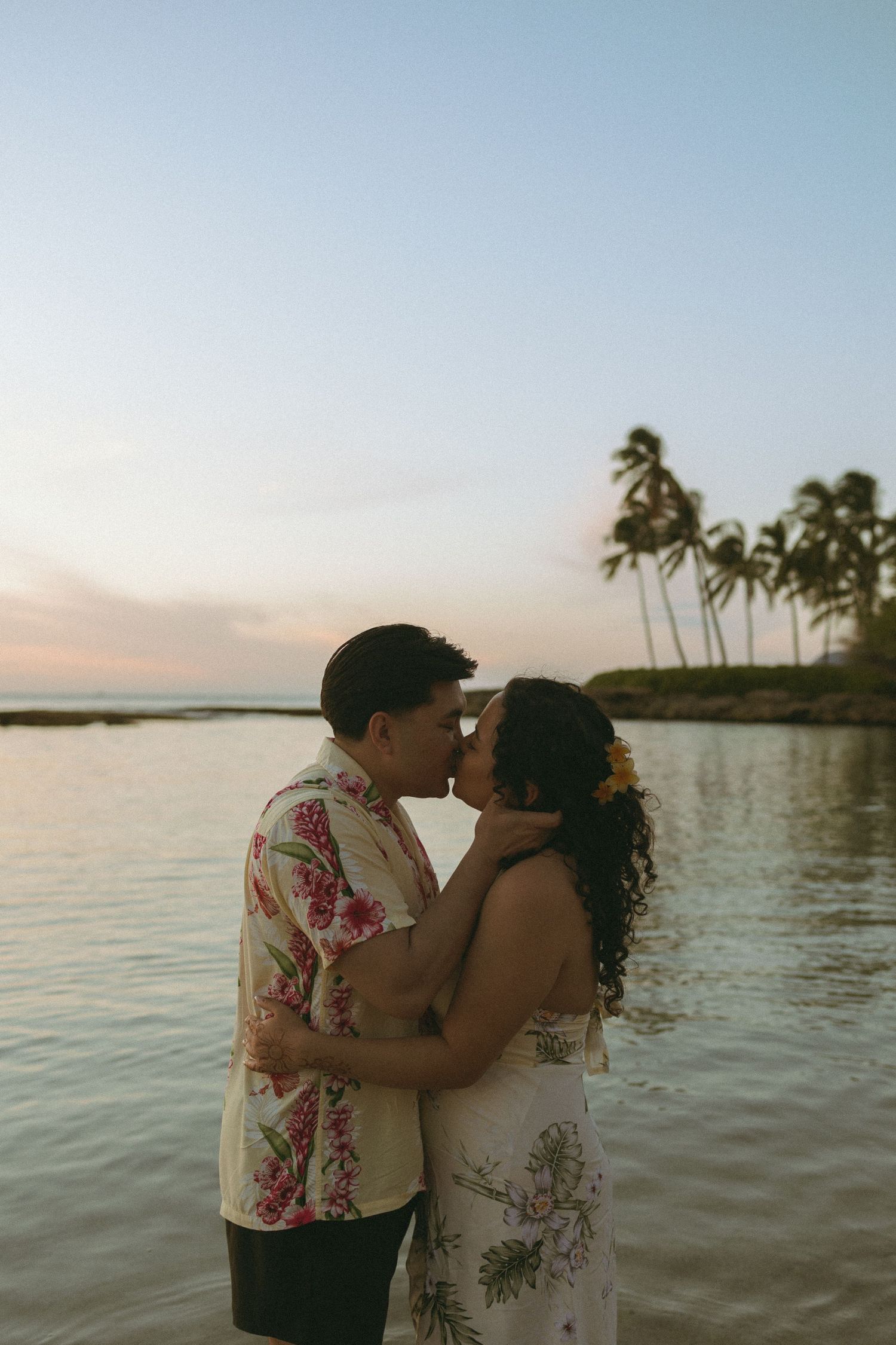 Couple sharing a romantic kiss at sunset on a Hawaiian beach, with palm trees and calm ocean waters in the background.