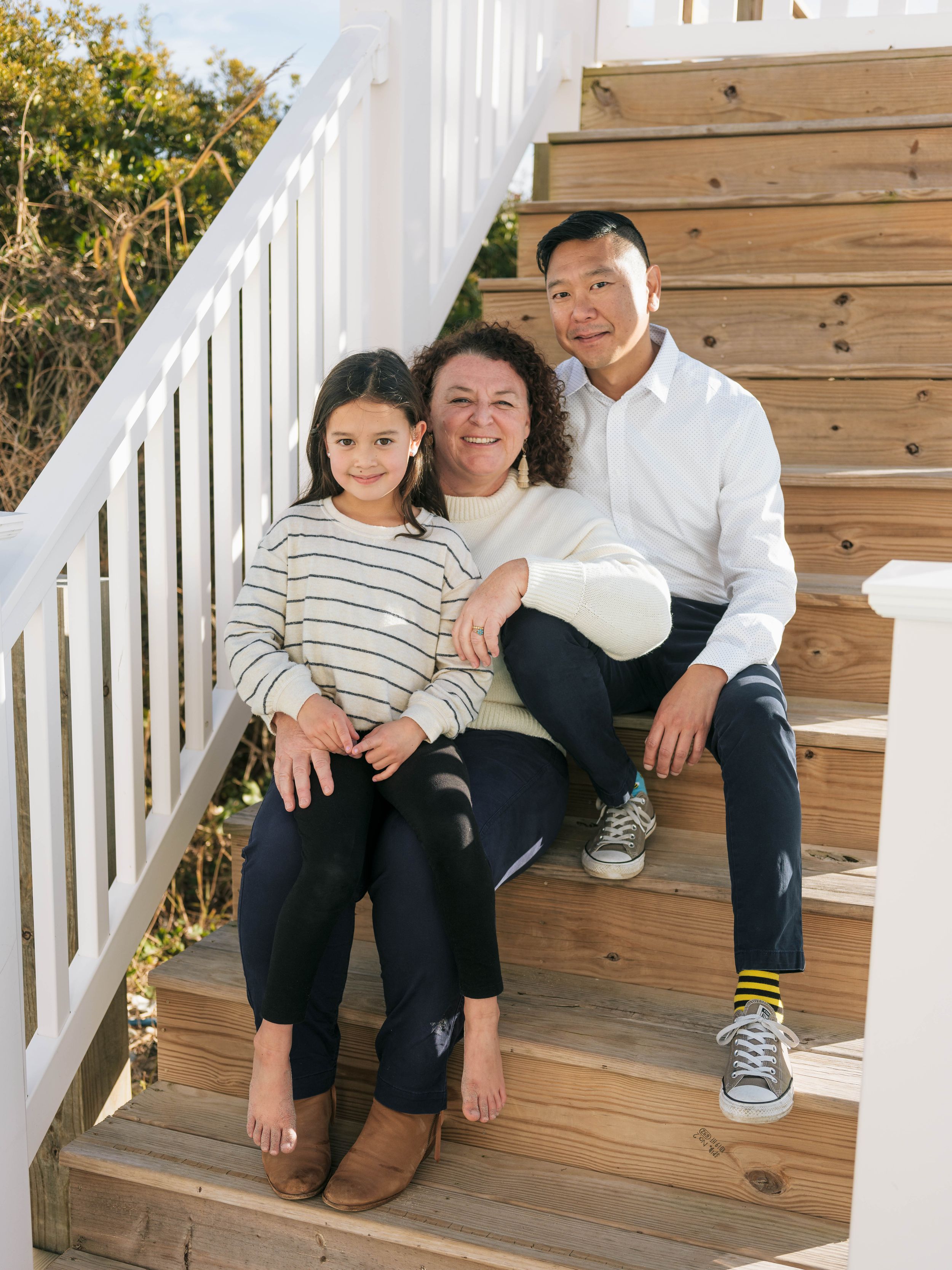 A family of three poses together on wooden outdoor stairs, smiling warmly in casual attire on a sunny day.