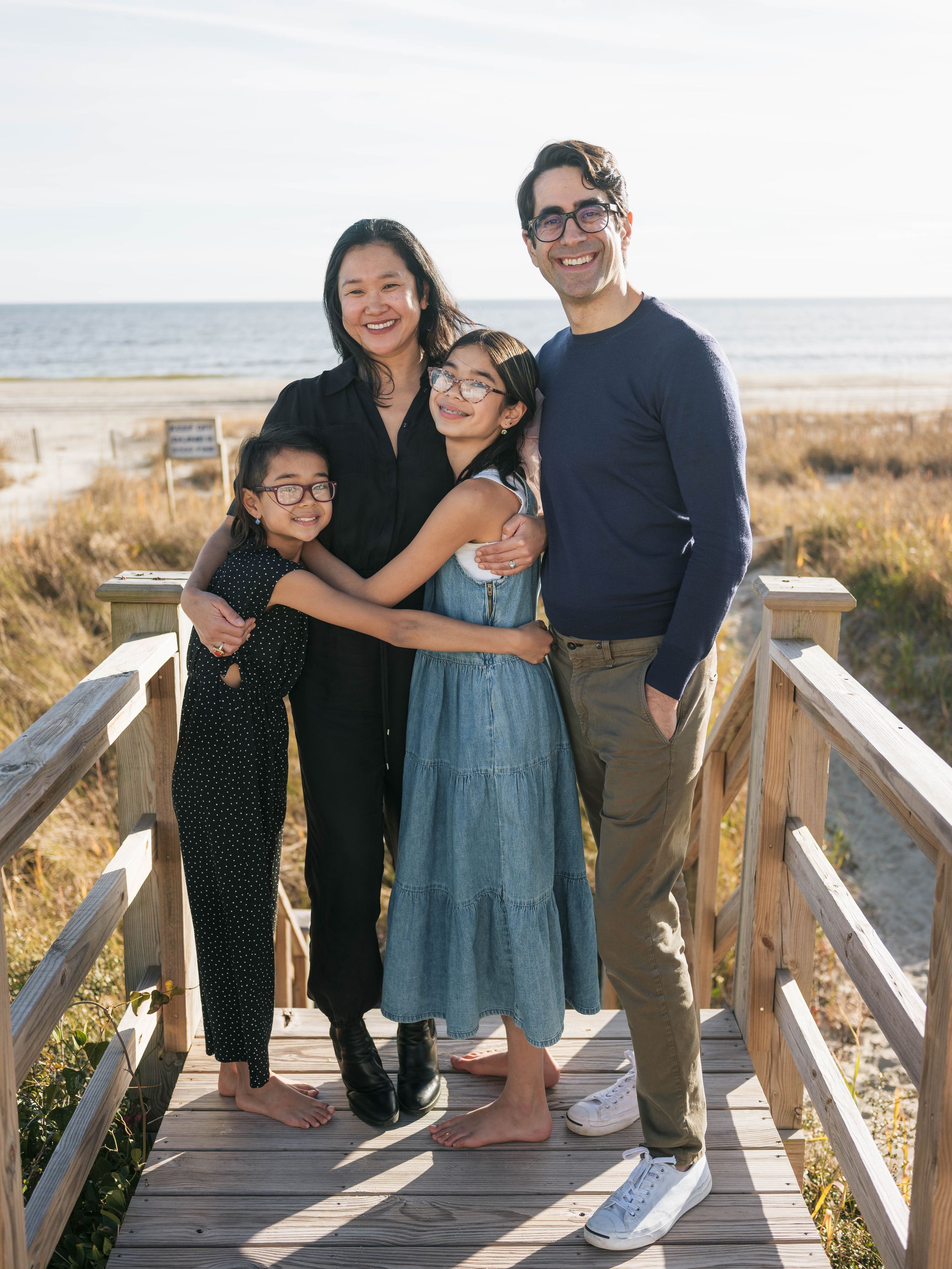 Family of four smiling together on a beachside boardwalk on a sunny day.