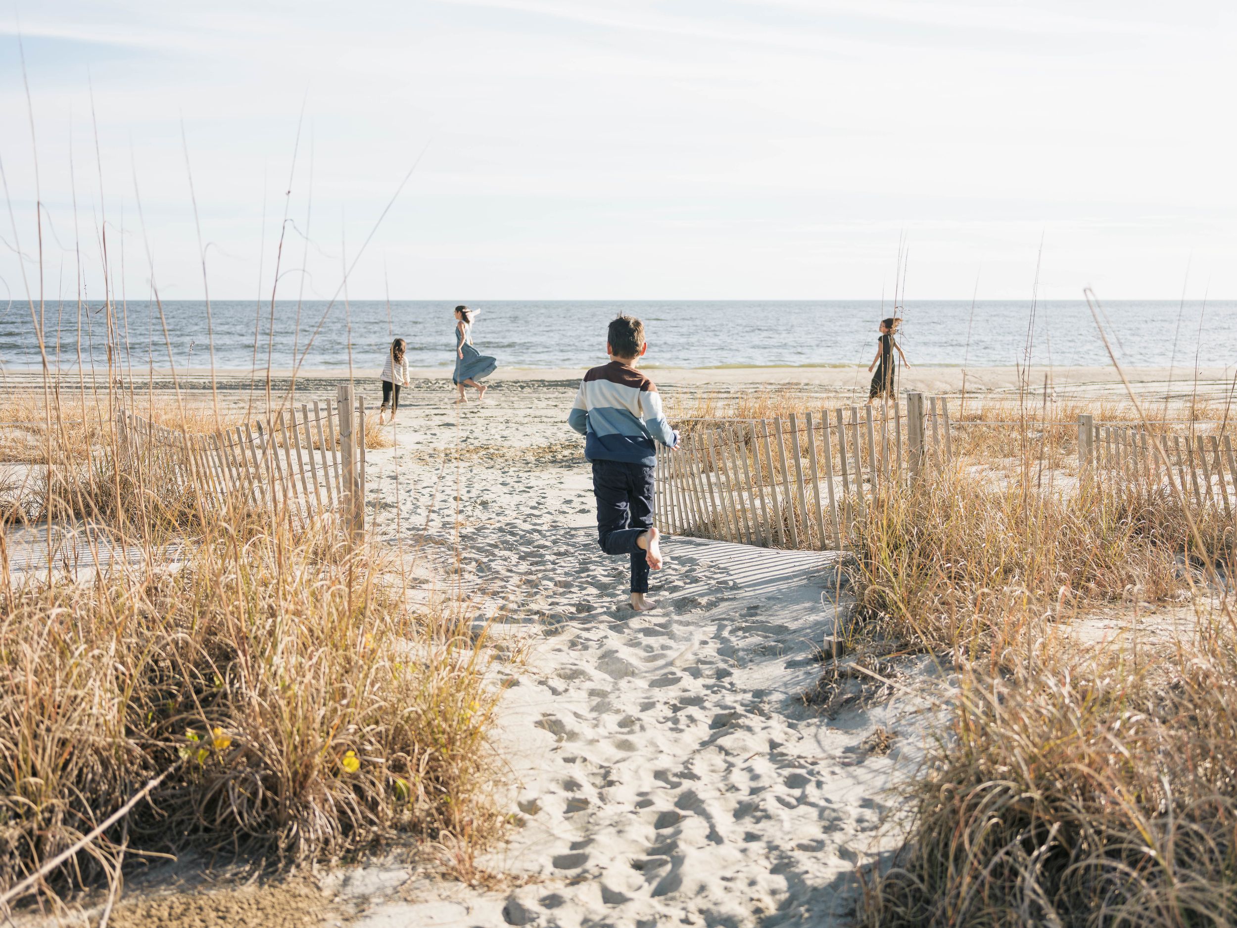 Child running down a sandy beach path toward the ocean, flanked by dune grass and wooden fencing.
