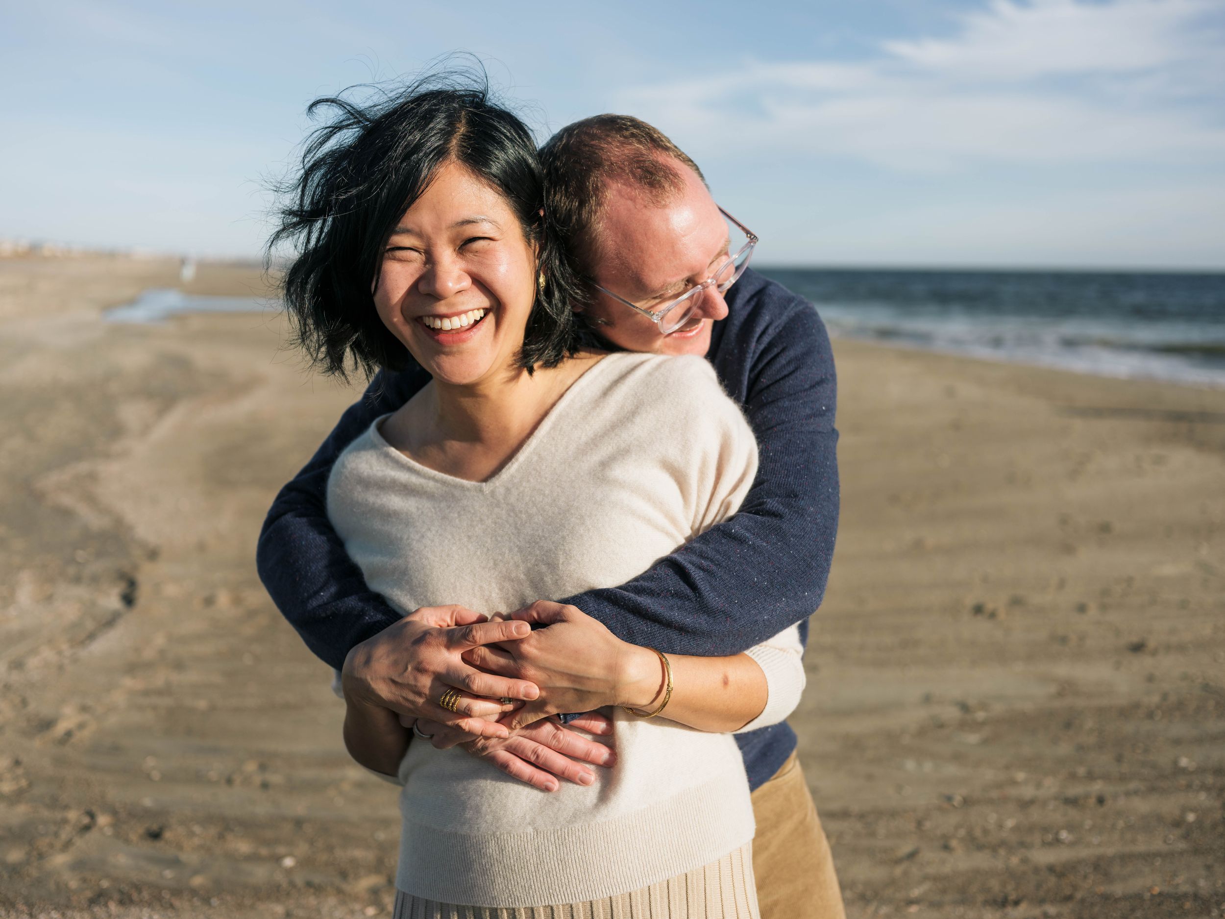 Happy couple embracing on a sandy beach, woman laughing joyfully while man hugs her from behind on a sunny day.