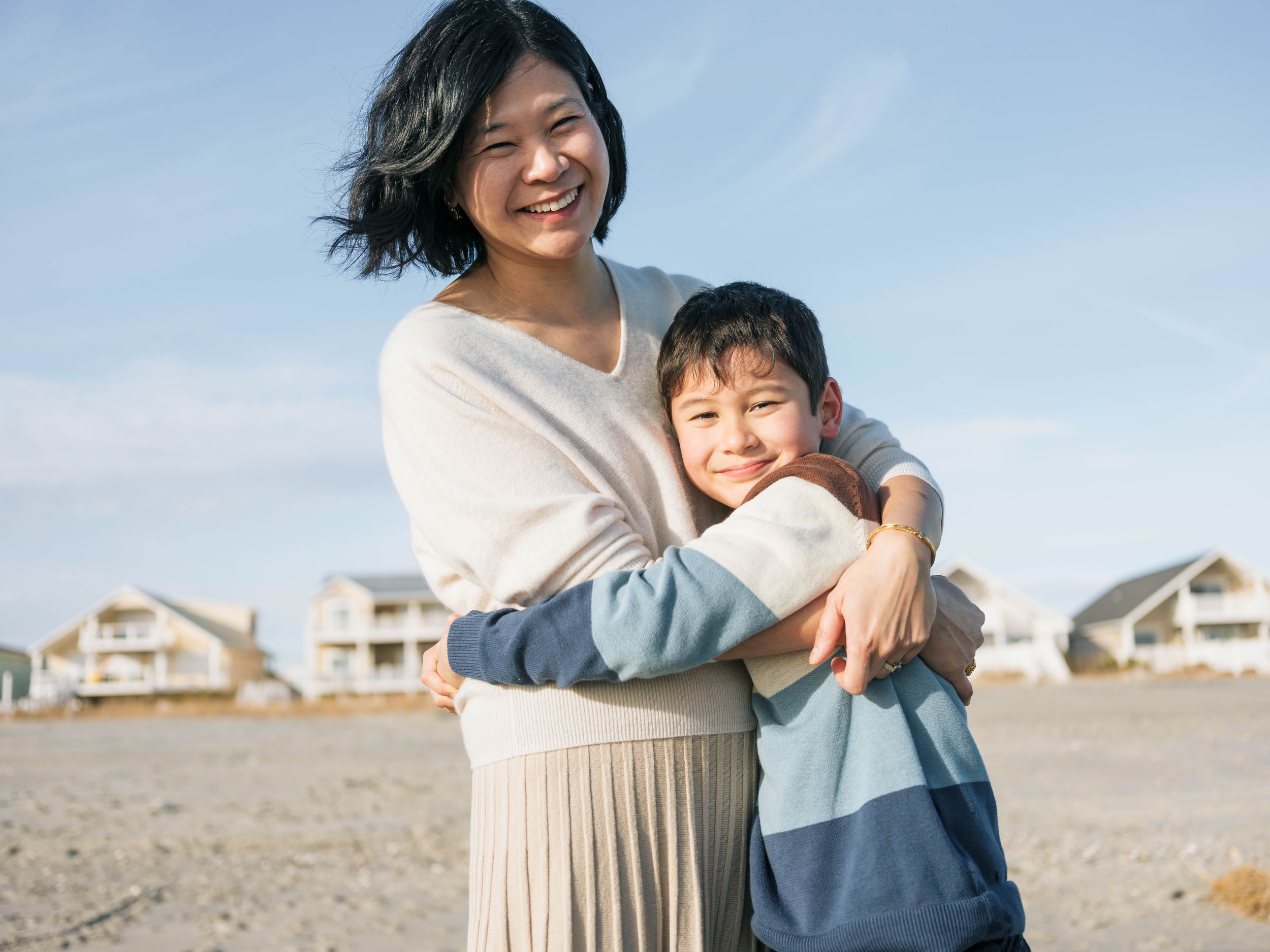A smiling Asian mother and young son share a warm hug on a sandy beach with houses in the background.