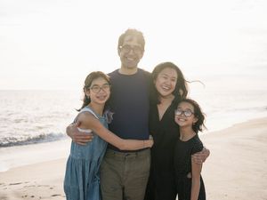 Family of four smiling together on a sunny beach, with ocean waves in the background.