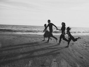 Black and white photo of three children running joyfully along a sandy beach with ocean waves in the background.