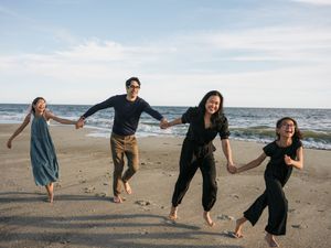 Family of four holding hands and jumping joyfully on a sandy beach with ocean waves in the background.
