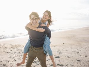A happy couple enjoys a piggyback ride on a sunny beach, smiling and laughing together.