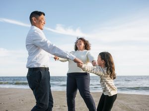 Family of three holding hands and spinning together on a sunny beach with ocean views.
