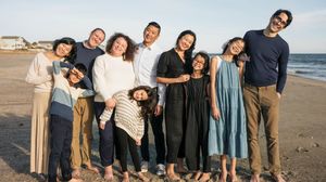 Large diverse family of nine smiling together on a sandy beach at sunset in casual clothing.