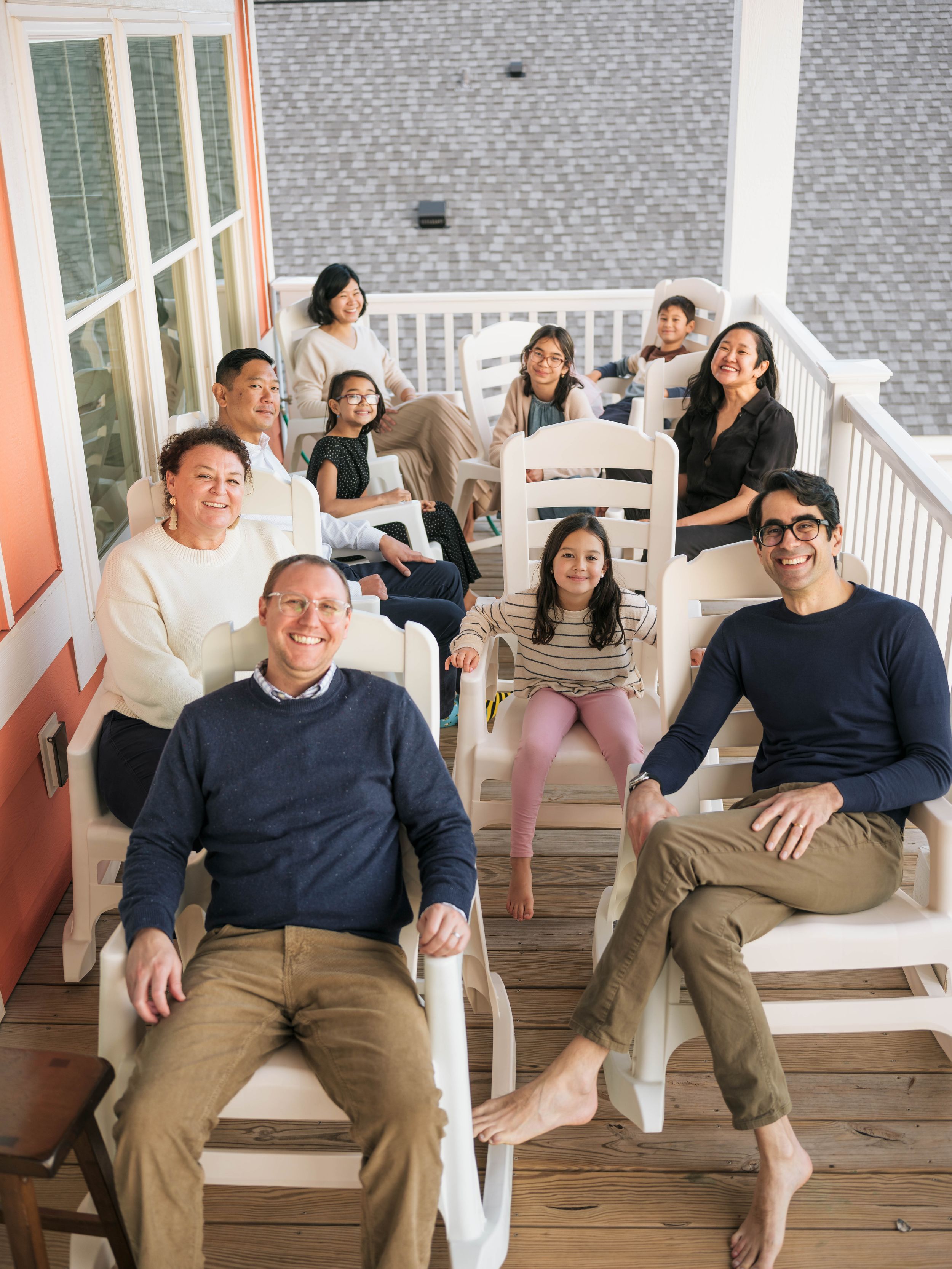 A multigenerational family smiles together on a porch, seated in white chairs during a casual outdoor portrait.
