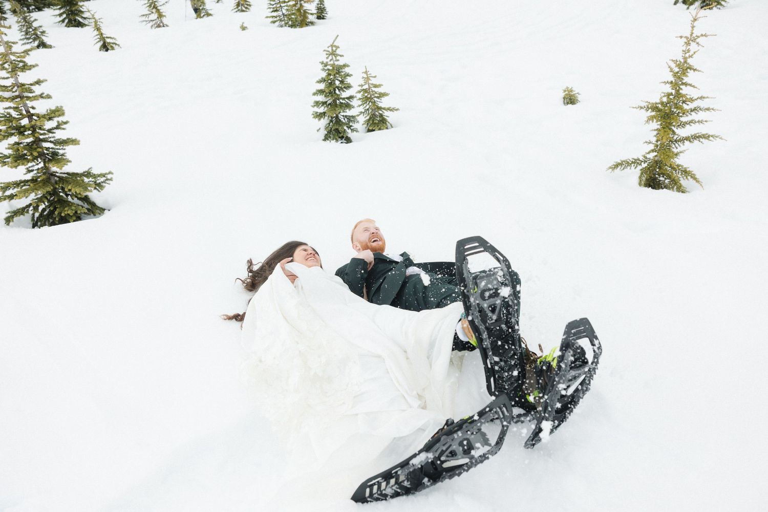 Couple in wedding attire playfully falling in snow wearing snowshoes surrounded by snow-covered evergreen trees.