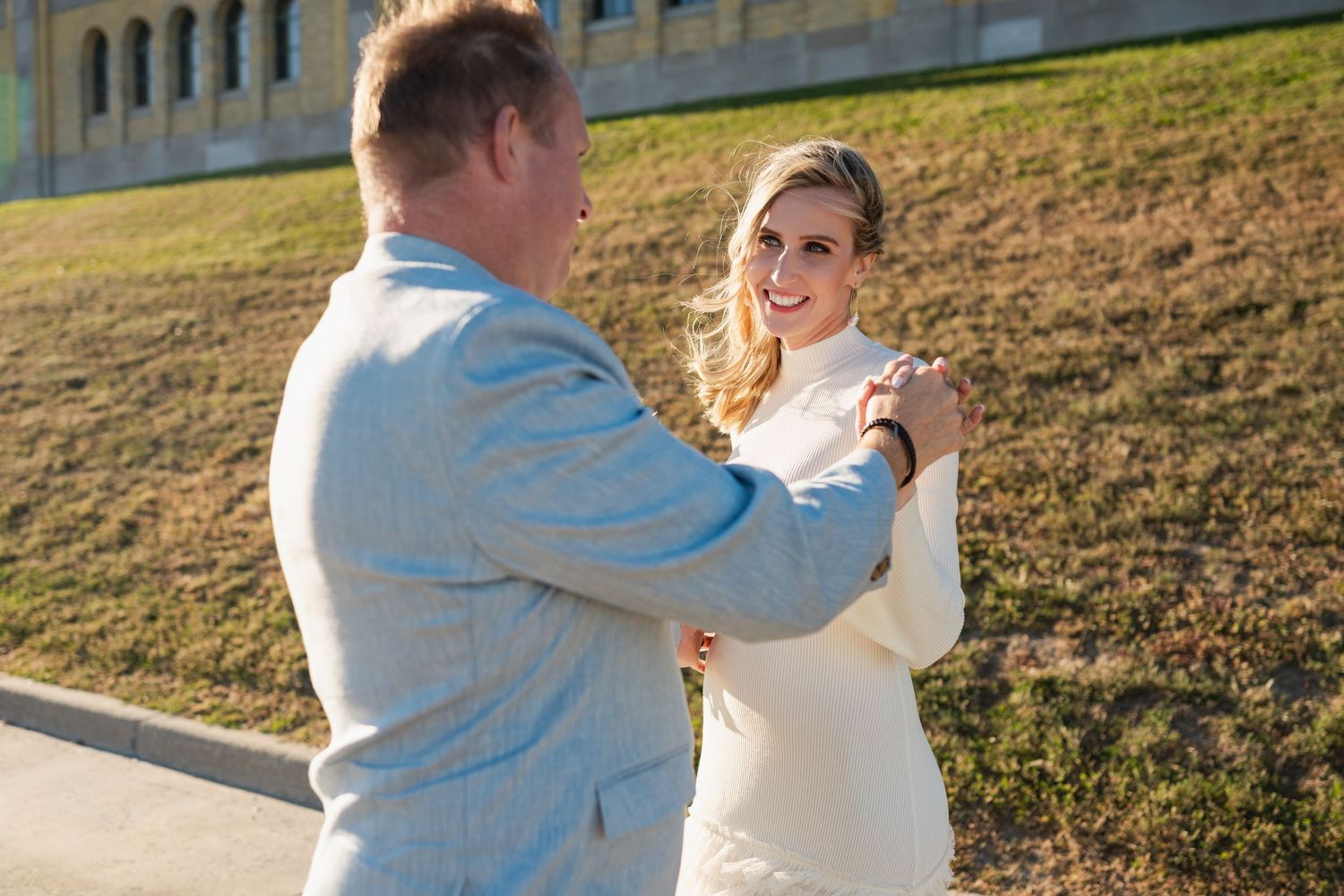 Bride and groom walking hand-in-hand on a grassy coastal hill with a historic building and ocean in the background.
