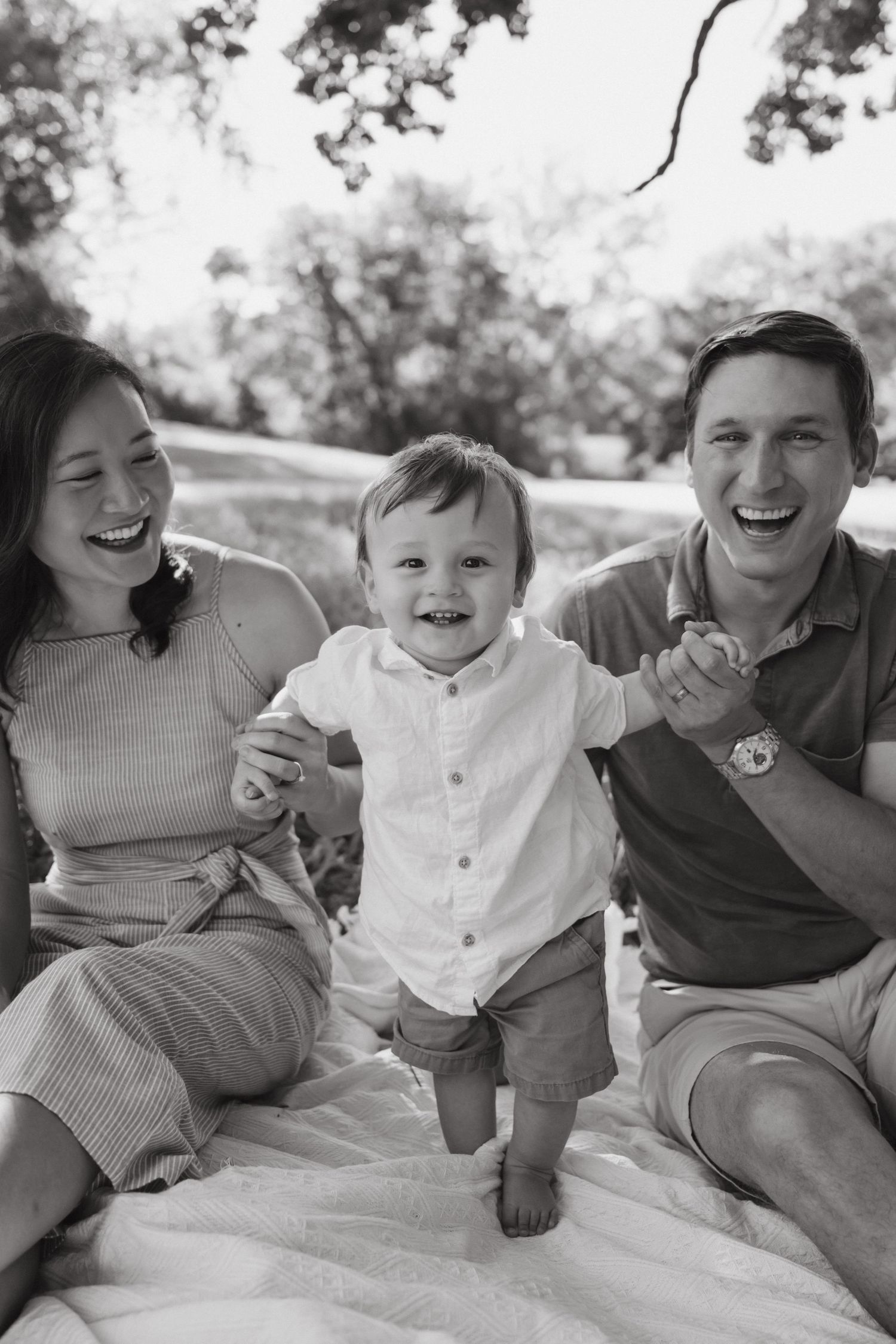 Black and white photo of happy parents laughing with their toddler son outdoors on a blanket in a park.