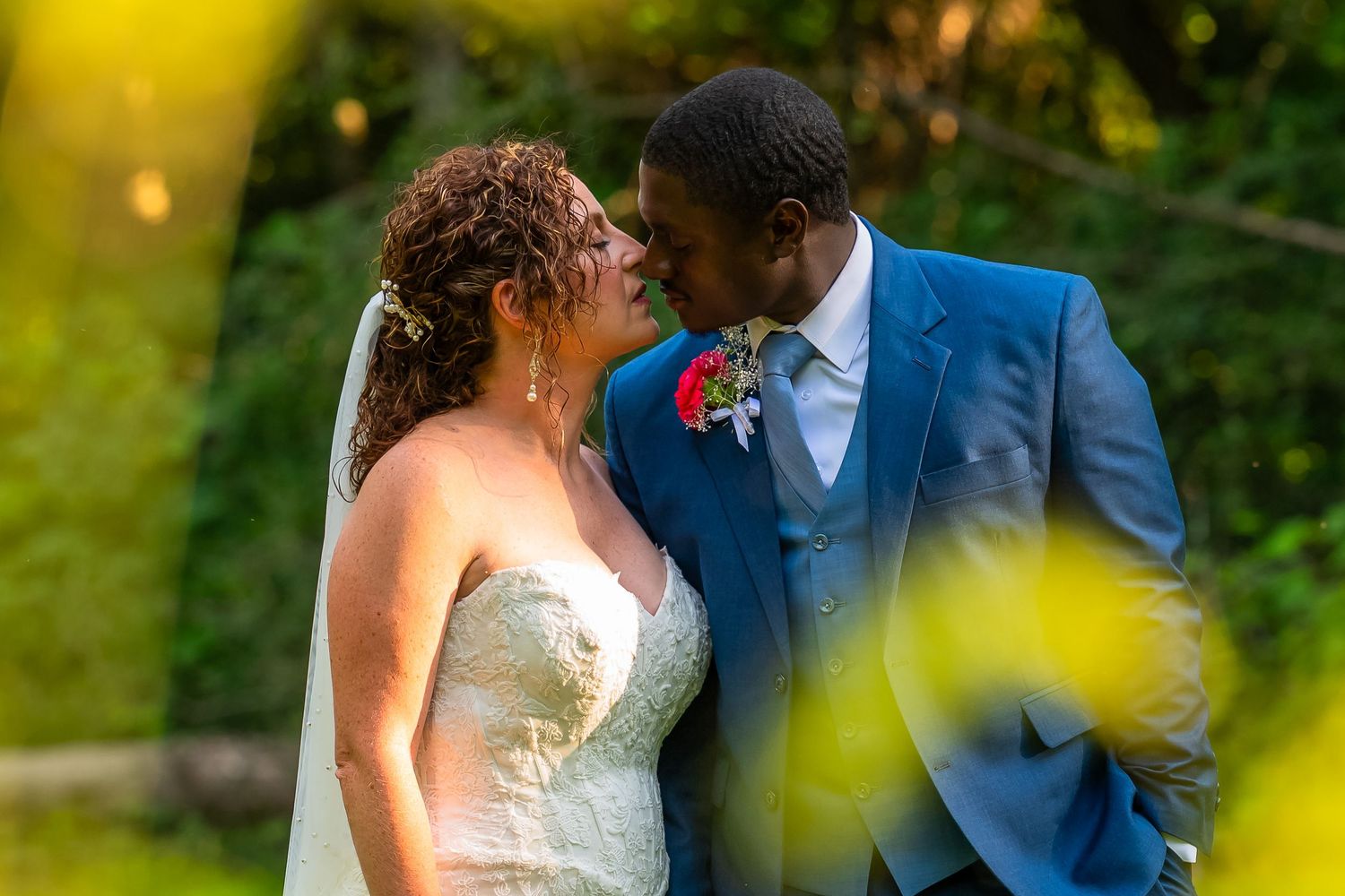 A bride and groom share a romantic almost-kiss outdoors, with the groom in a blue suit and bride in a white lace gown.