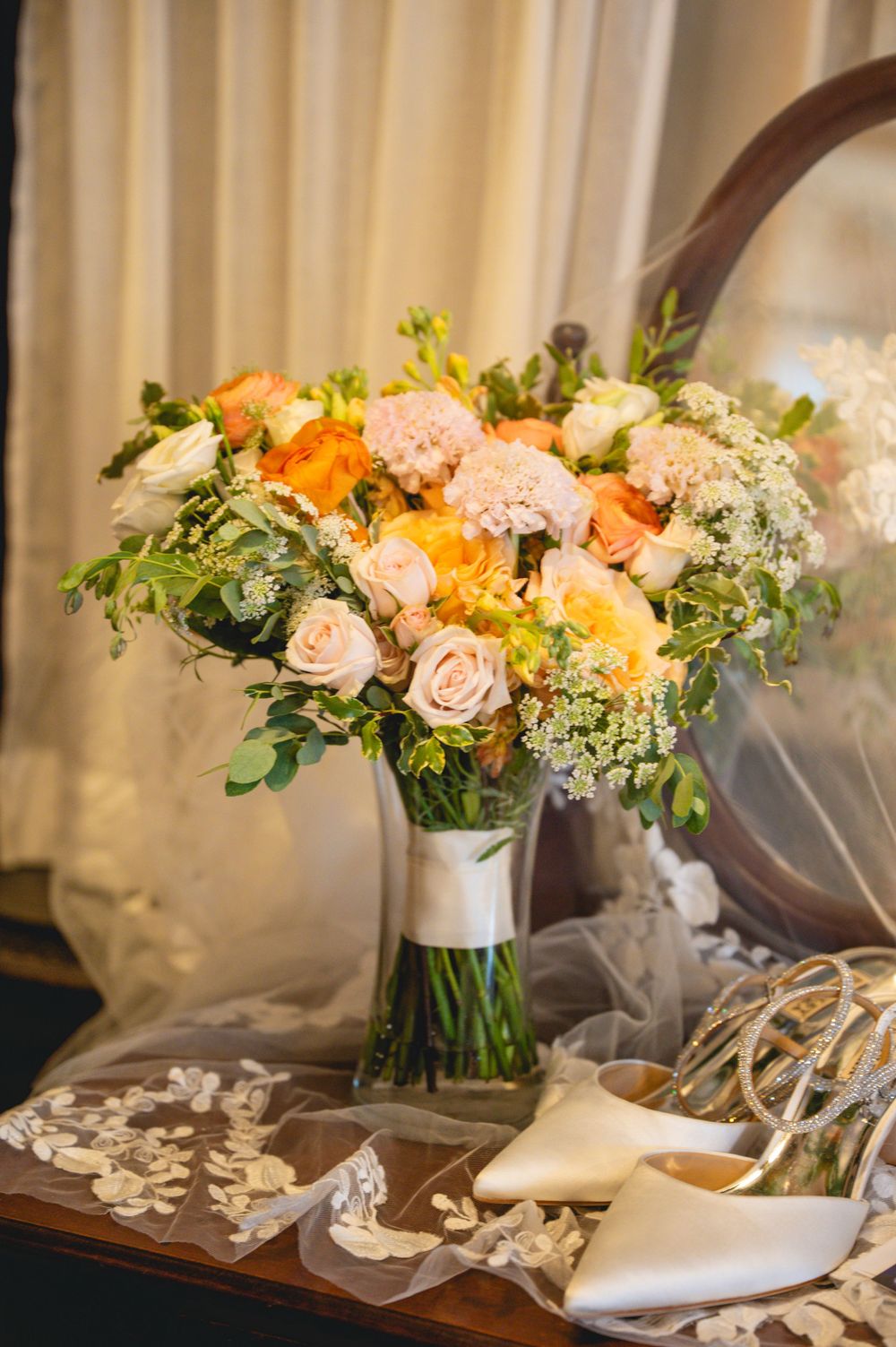 Elegant wedding bouquet of peach and orange roses in a vase beside bridal shoes and lace veil on a wooden table.