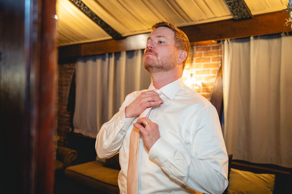 Groom adjusting his tie while getting ready in a warmly lit rustic room before the wedding ceremony.