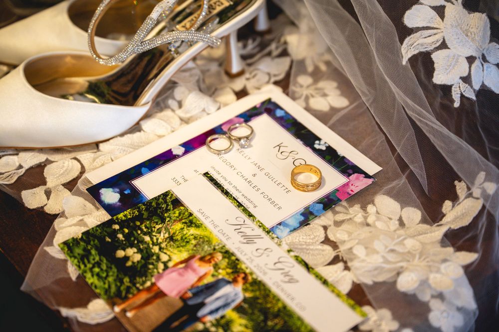 Wedding flat lay with white heels, gold rings, floral veil, and elegant invitation cards on a decorative surface.
