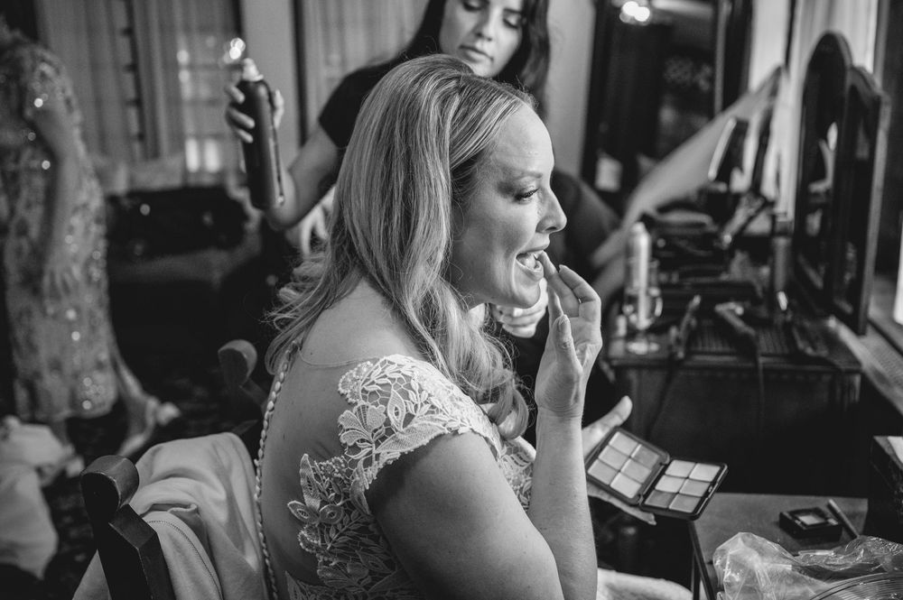Bride applying lipstick at vanity mirror while bridesmaid styles her hair in black and white wedding photo.