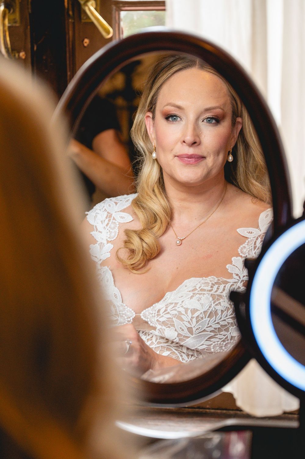 Bride in lace wedding dress gazes into oval mirror during bridal preparations, with soft glam makeup and elegant jewelry.