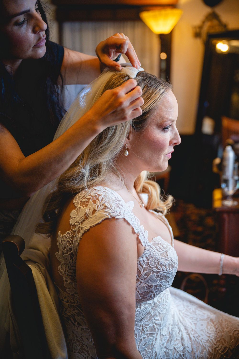Bride in lace wedding dress gets veil attached by stylist during bridal preparation before wedding ceremony.