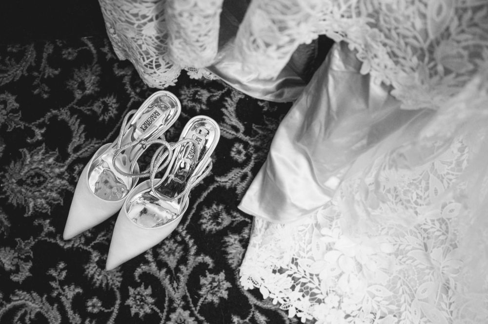 Black and white photo of elegant bridal heels beside a lace wedding dress on a patterned floor.