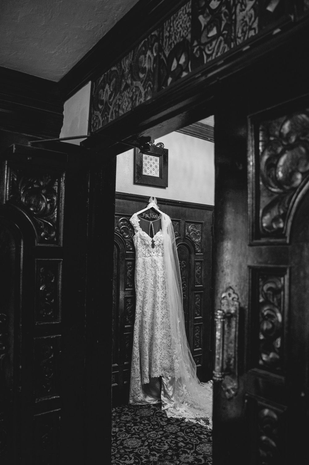 Black and white photo of an elegant lace wedding dress hanging in ornate dark wood doorway.