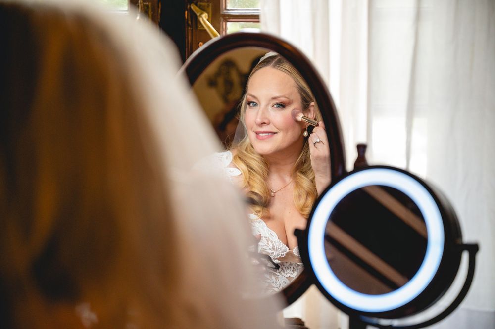Bride putting on earrings while looking into a lighted round mirror during wedding day preparations.