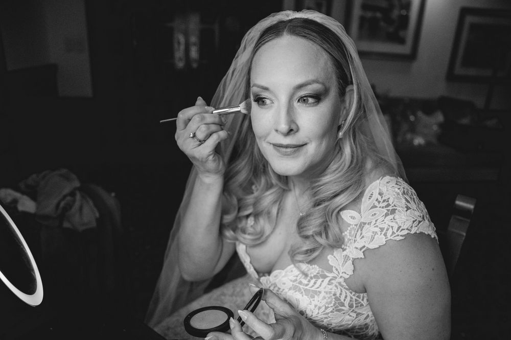 Bride in lace dress and veil applying makeup with brush in black and white wedding preparation photo.