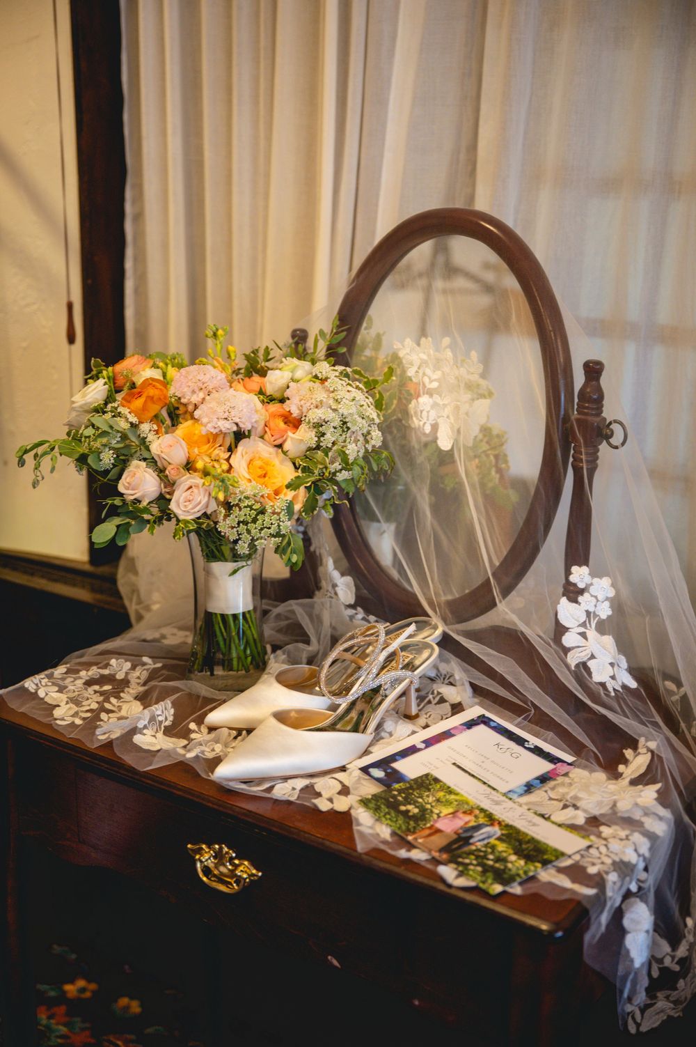 Elegant bridal vanity with oval mirror, white heels, lace veil, and a bouquet of peach and pink roses on a wooden dresser.