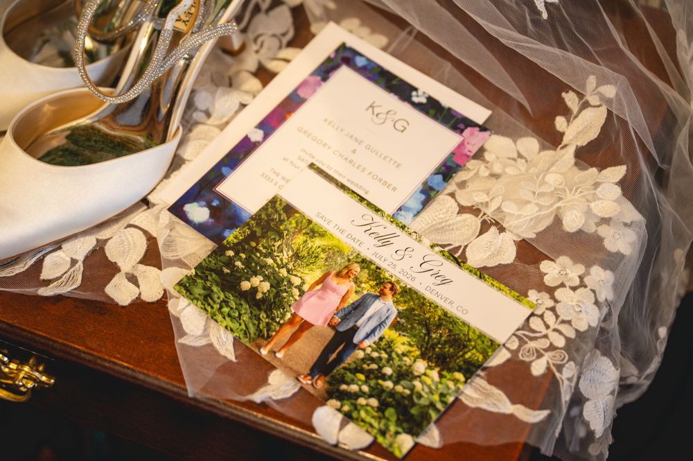 Wedding details flat lay featuring invitation suite, couple photo, bridal veil, and heels on wooden tray.