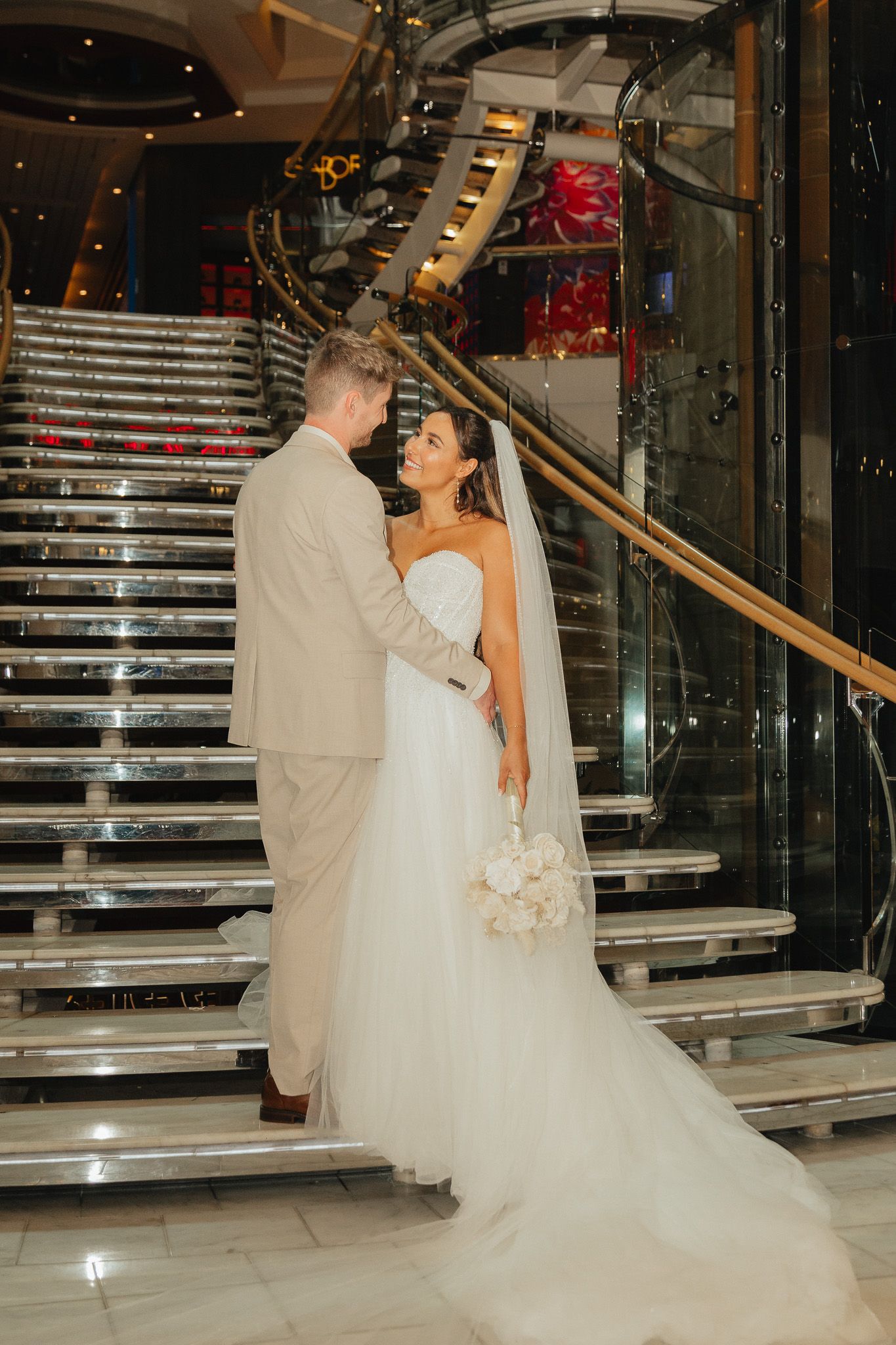Bride and groom share a romantic moment on a grand staircase, bride in white gown holding bouquet.