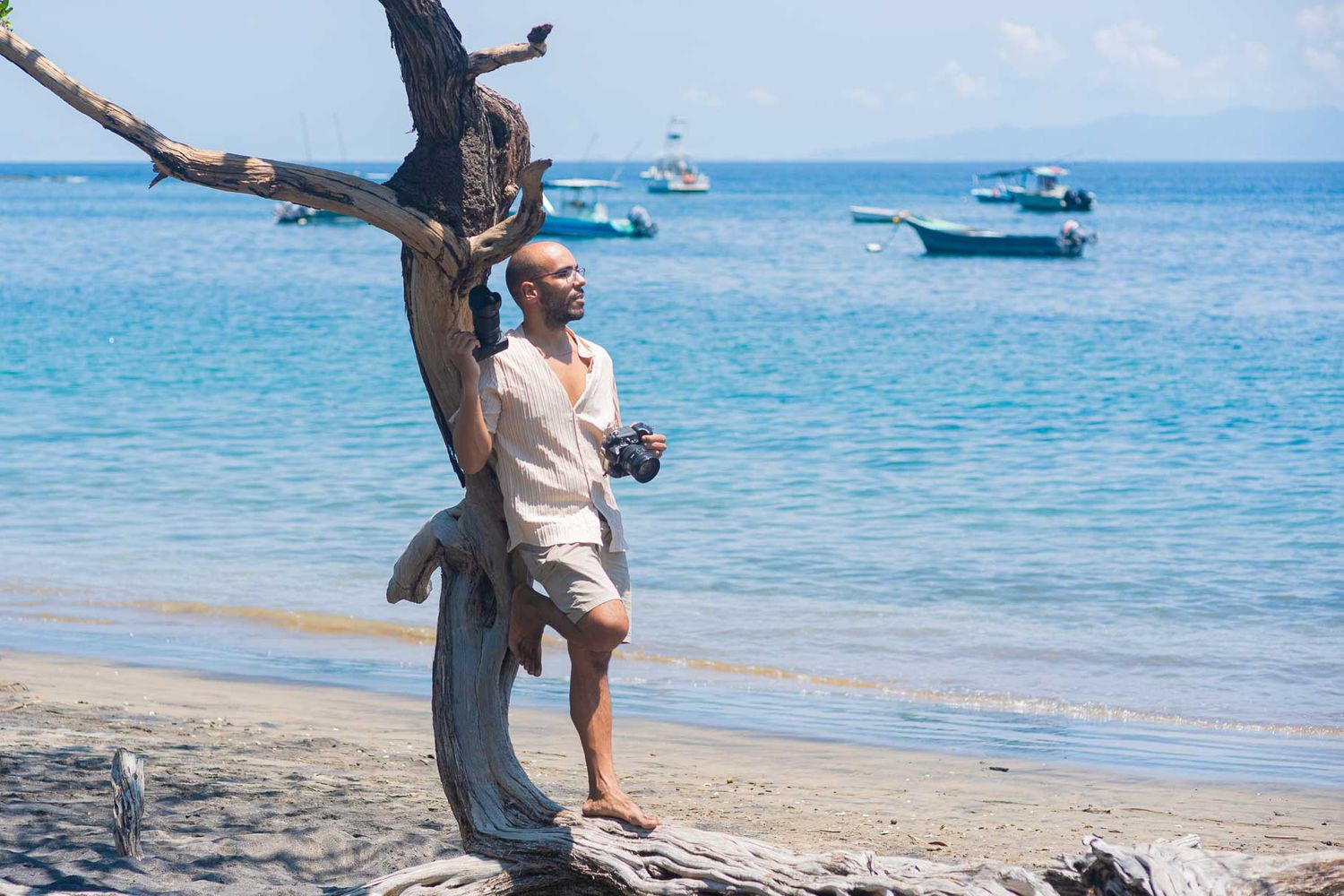 A bald man leans against a weathered tree on a tropical beach with turquoise water and boats in the background.