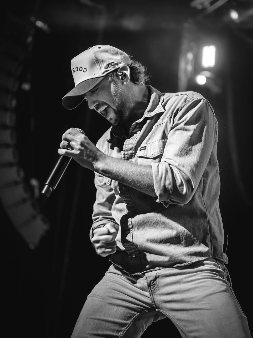 Black and white photo of a male singer performing on stage, holding a microphone and wearing a cap and button-up shirt.