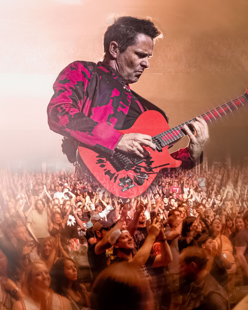Guitarist performing on stage with red electric guitar over large concert crowd in dramatic lighting.
