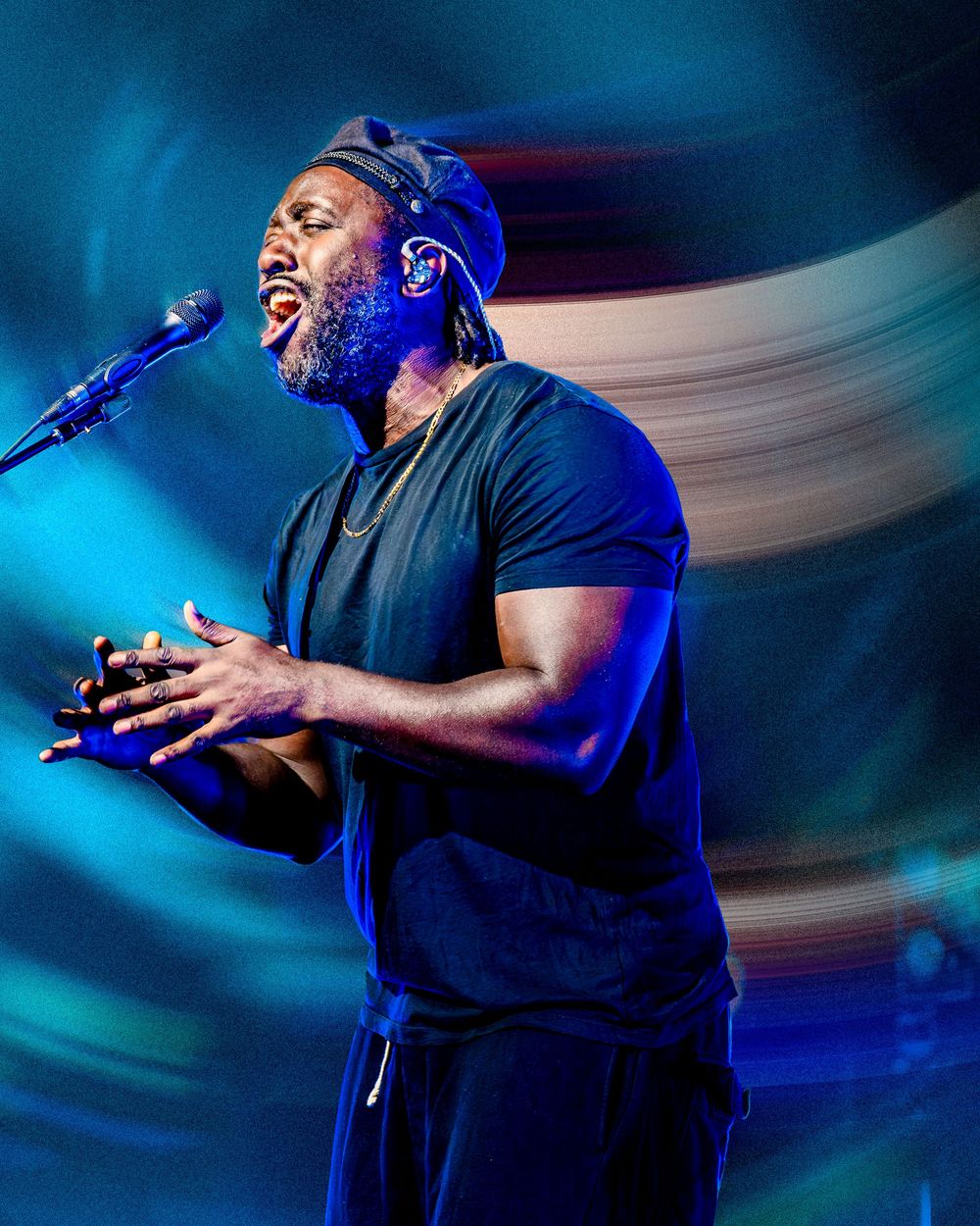 A Black male singer performs passionately on stage, wearing a black t-shirt and cap, with blue and white swirling lights behind him.