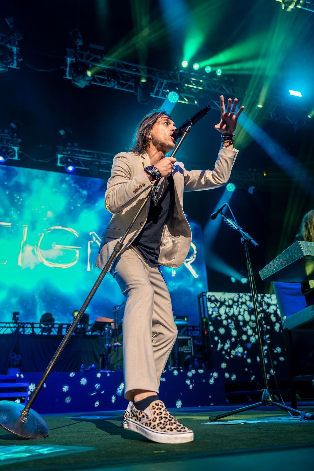 Energetic male performer sings on stage with dramatic blue and green lighting at live concert.
