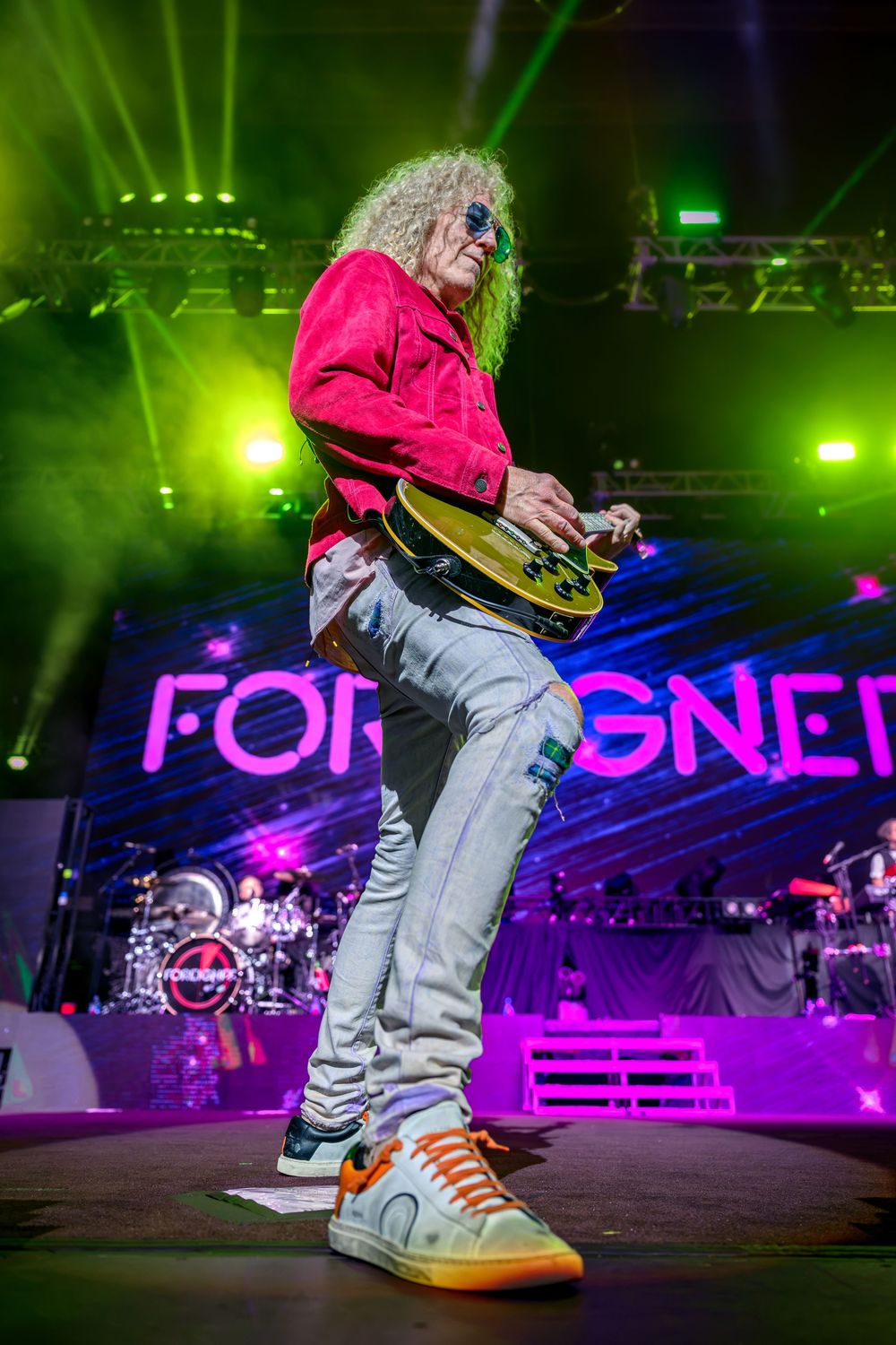 Guitarist performs on stage at a Foreigner concert, wearing a pink jacket and jeans under vibrant green lights.