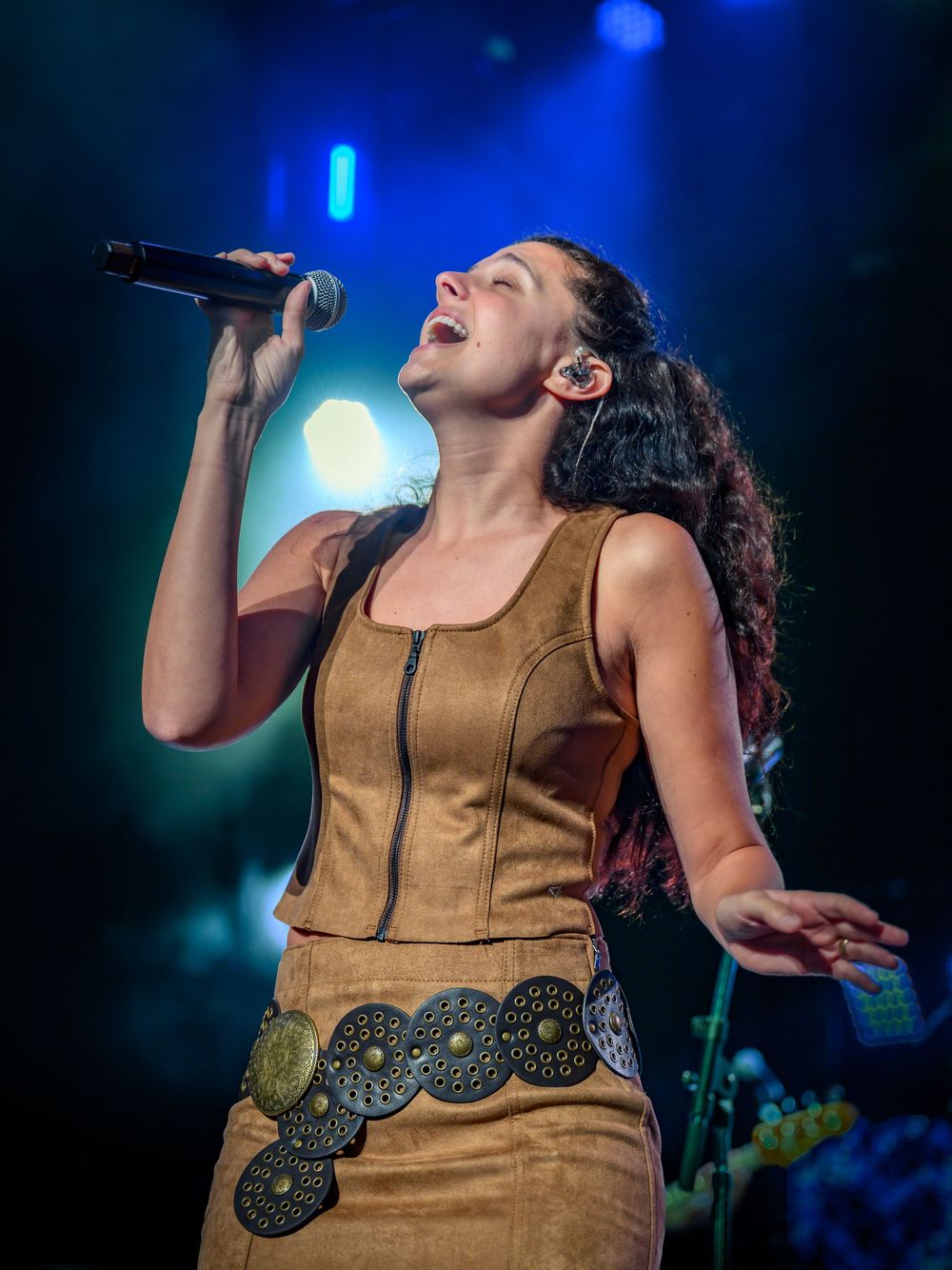 A female singer performs passionately on stage, holding a microphone, wearing a tan suede outfit with decorative medallion belt.